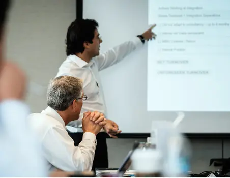 Twee personen in een vergaderruimte, waarvan één wijst naar een projectiescherm met tekst over softwaretechnologie en klantondersteuning.