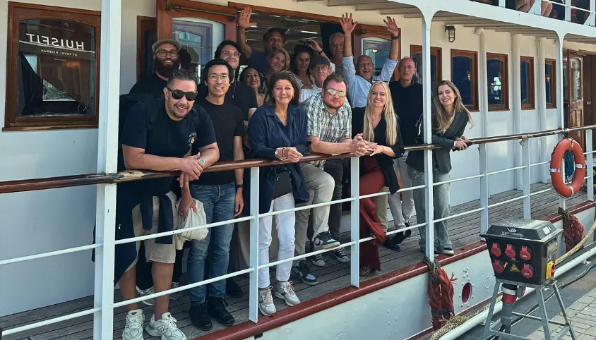 Groep mensen poseert op het dek van een boot met witte relingen en houten accenten; sommigen hebben hun handen in de lucht.