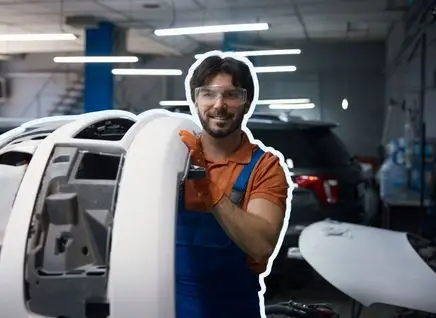 A person in an auto workshop working on a large bodywork component, with various vehicles and tools visible in the background.