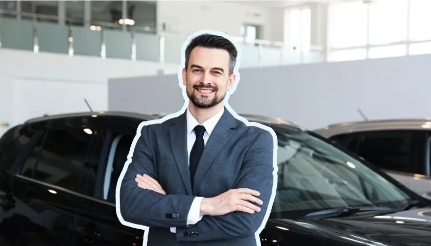 A person in a neat business outfit standing with crossed arms in front of a car inside a modern showroom.